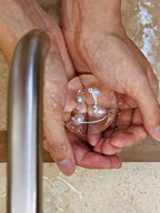 Hands holding a transparent handmade hydrating soap with four genuine freshwater pearls under running water.